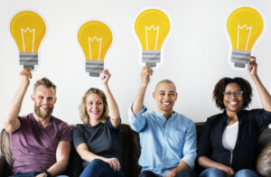 A group of diverse team members hold a large cutout of a light bulb over their heads to illustrate the ideas that can be generated when engaged in an inspired team.